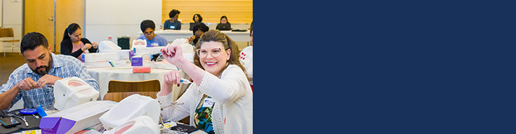 Person attending a video conference on a laptop, where a woman is presenting with a clipboard to a group of participants.