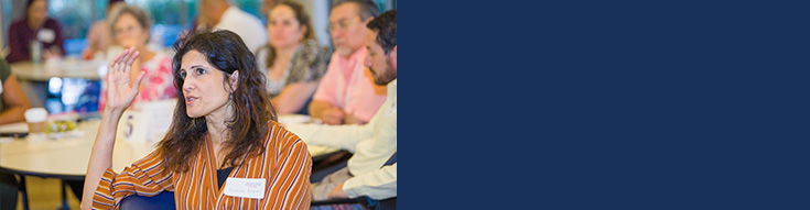 Male doctor in a white coat sitting at a desk, writing in a notebook while looking at a laptop screen during a virtual consultation.
